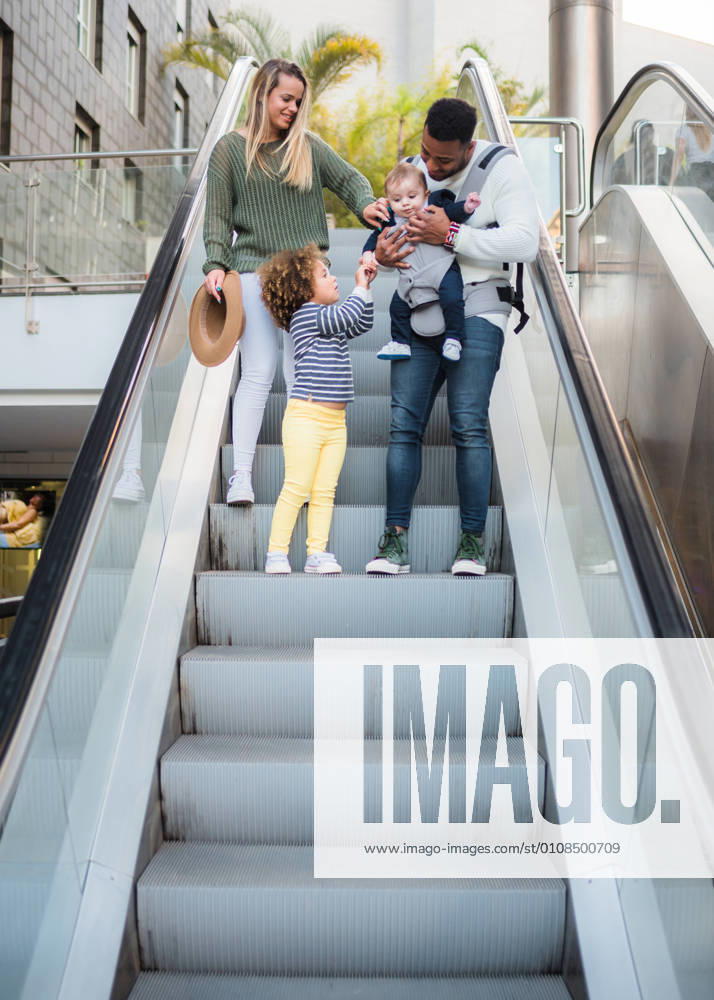 From below of happy multiracial family going downstairs on escalator and little girl taking care of baby brother carried by dad while spending together summer day in city, Model released