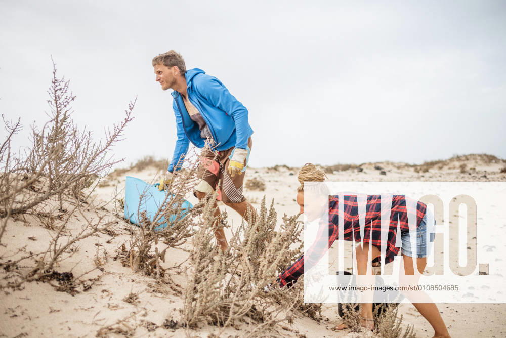 IMAGO Creative: N?chste Generation, Sozial- und klimabewusst Positive hipster woman and man in casual clothes and gloves collecting trash into bag while squatting on deserted beach
