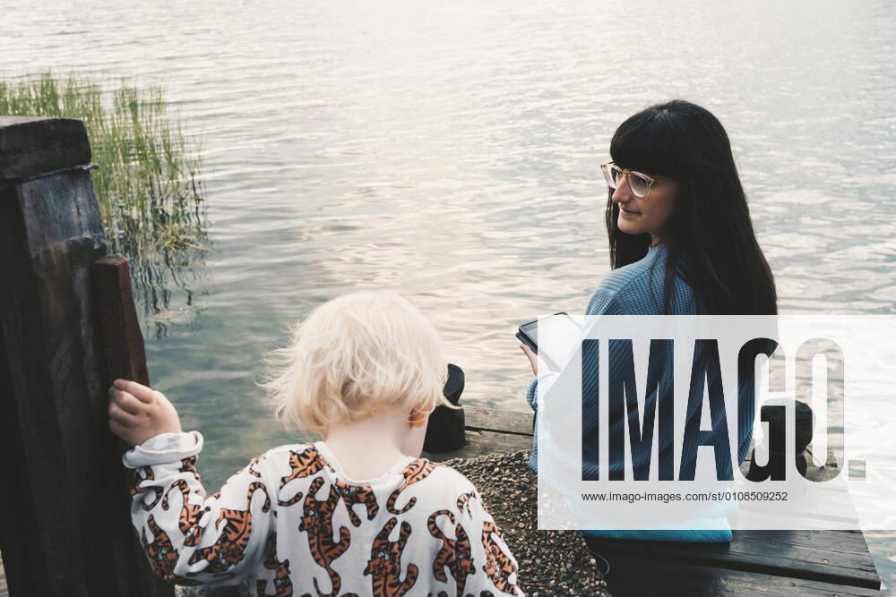 Mother looking at daughter while holding digital tablet and sitting on jetty over lake , model released, property released