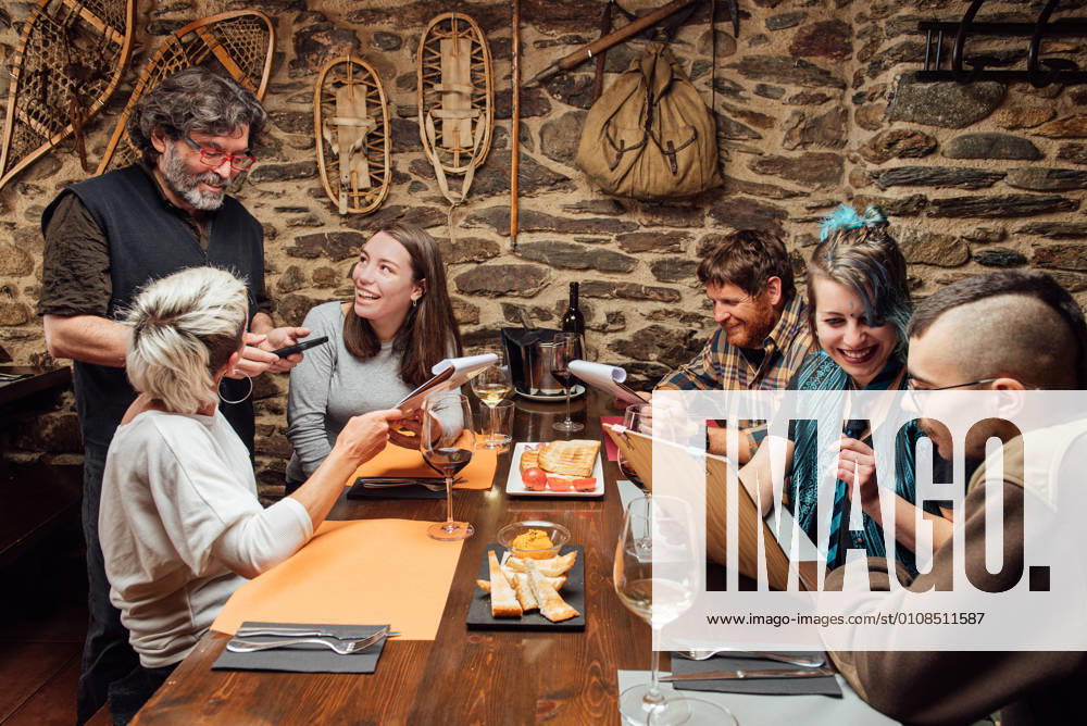 IMAGO Creative: Landleben - Essen Cheerful group of hipsters sitting at wooden table with menu and talking to waiter while ordering food during date in cafe, Model released, Property released