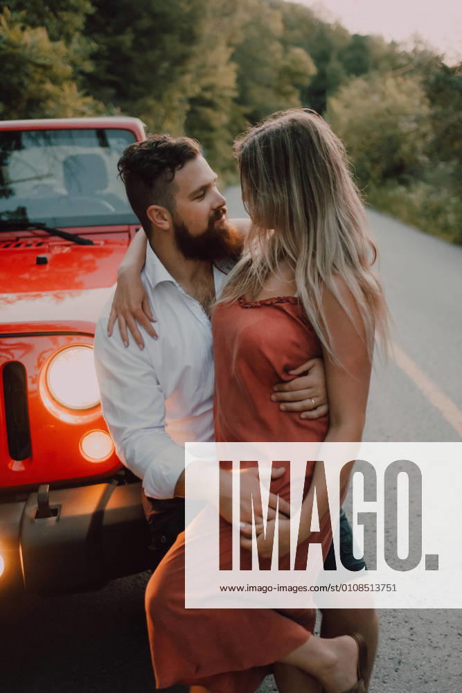 Tender bearded man in white shirt and black shorts hugging smiling woman in dress while sitting on red car hood on highway near green forest
