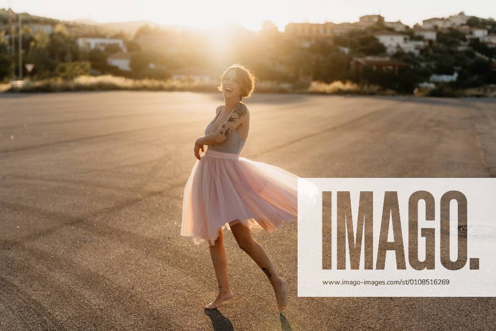 IMAGO Creative: Lokalhelden - Kunst und Kultur Full body delighted young female artist in elegant dance dress laughing and dancing in back lit in empty square with blurred town in background in sunny summer sunset day