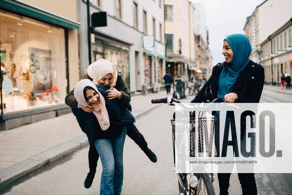 Young woman walking with bicycle looking at cheerful teenage girl giving piggyback to friend on street in city , model released