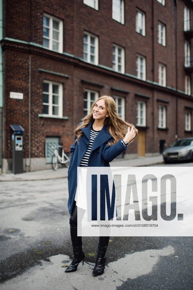 Full length portrait of smiling young woman standing with hand in hair on street against building in city , model released