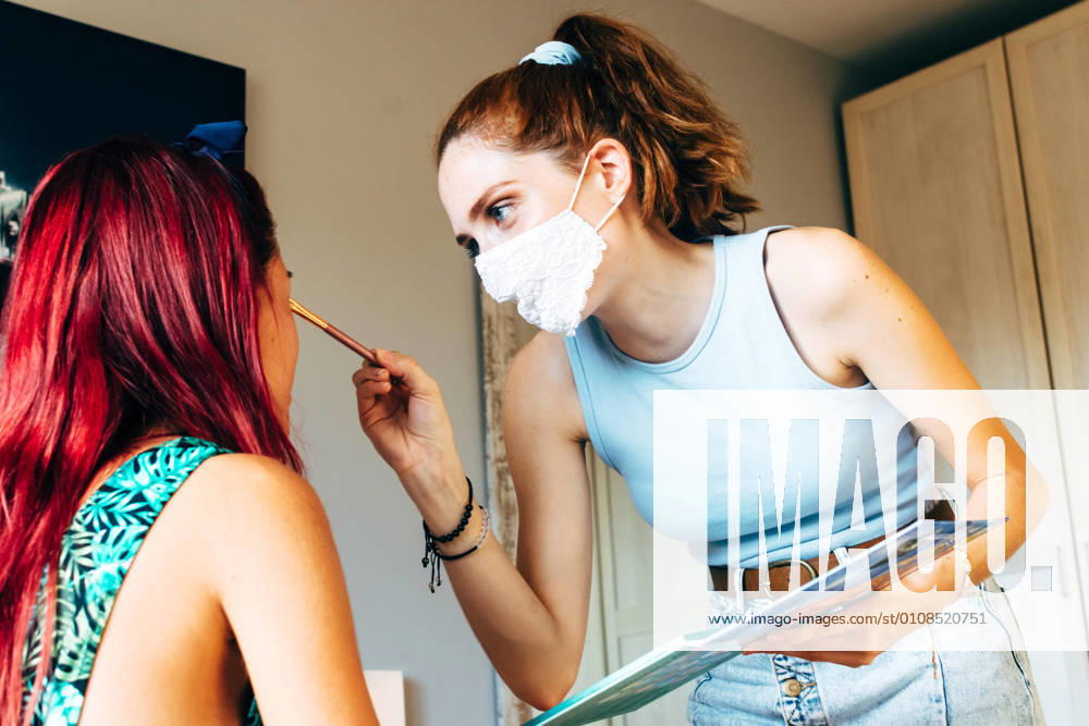 IMAGO Creative: Gesellschaft, Integration IMAGO Creative: Clickbait, Hautpflege und Kosmetik Side view of young female in casual clothes and protective mask with cosmetics palette and brush in hands doing makeup to girlfriend in bedroom at home, Model released