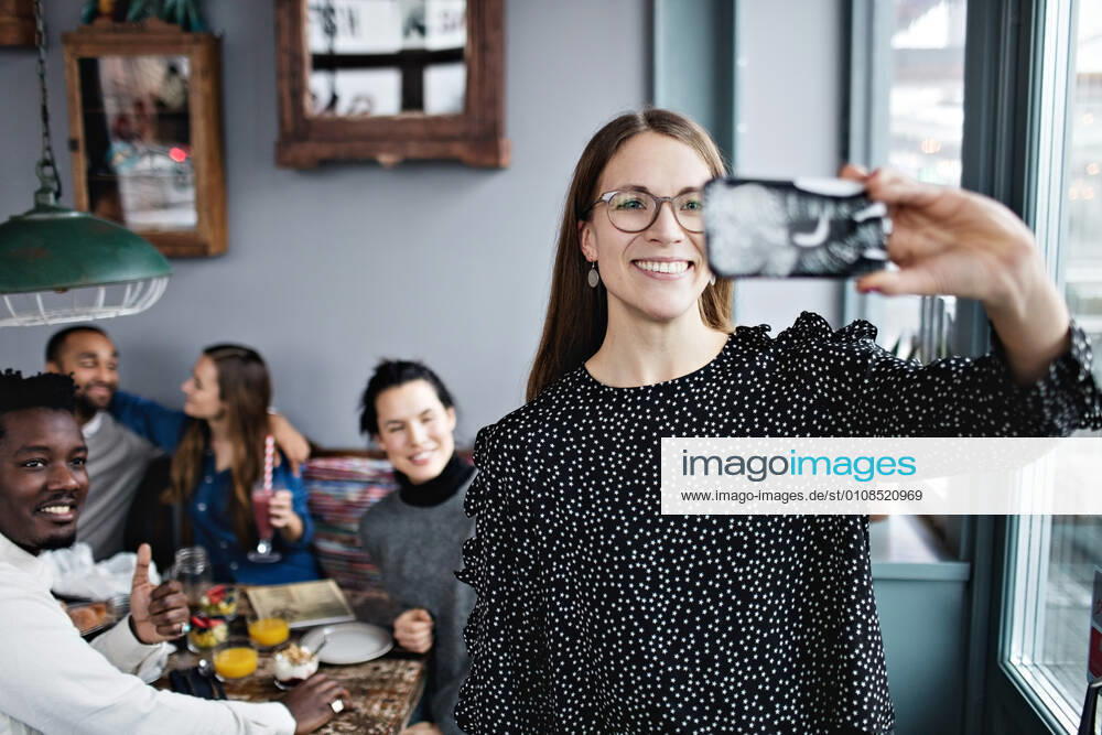 Smiling young woman taking selfie with friends sitting at restaurant , model released, property released