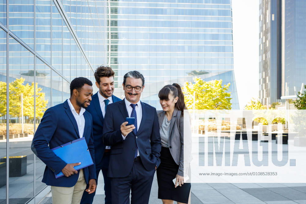 Group of happy multiracial colleagues in classic suites laughing while reading good news on smartphone standing together on street, Model released