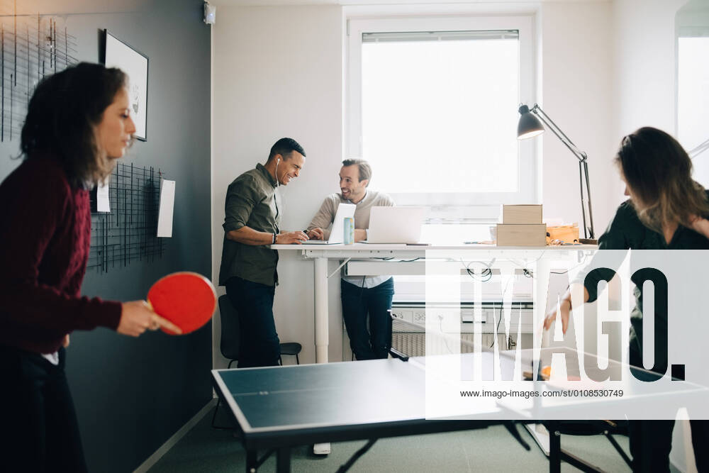 Female professionals playing table tennis while male colleagues using laptop at creative office , model released, property released