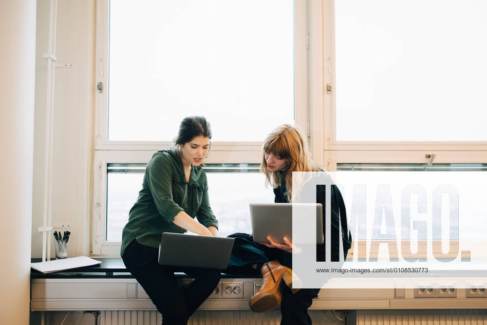 Confident female colleagues discussing over laptops while sitting on window sill at creative office , model released, property released