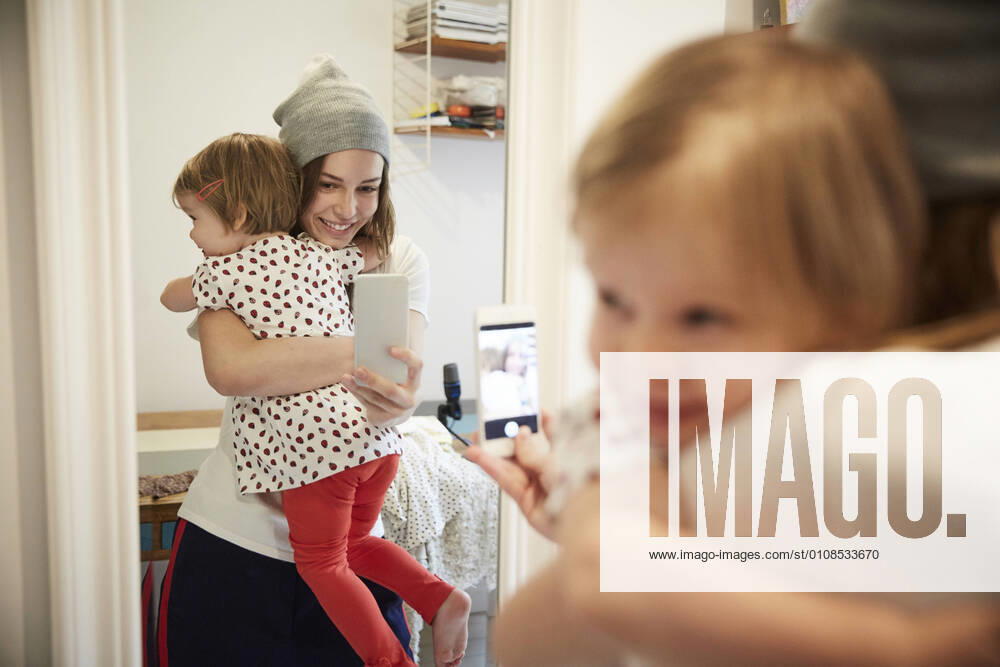 Smiling podcaster taking selfie with daughter through smart phone while reflecting on mirror , model released, property released