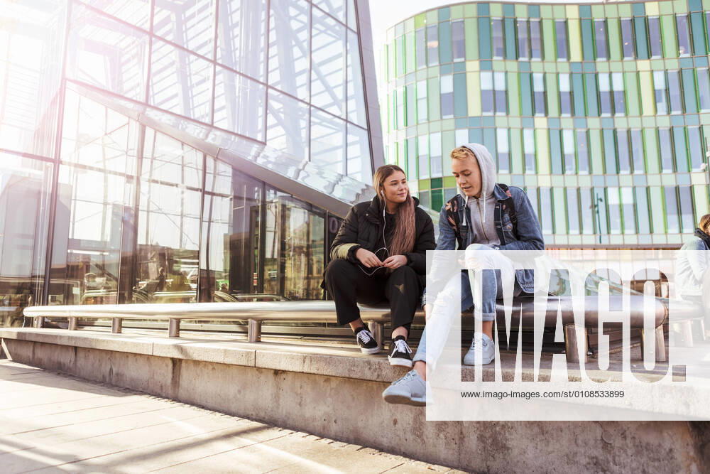Full length of teenage friends talking while sitting on railing in city , model released
