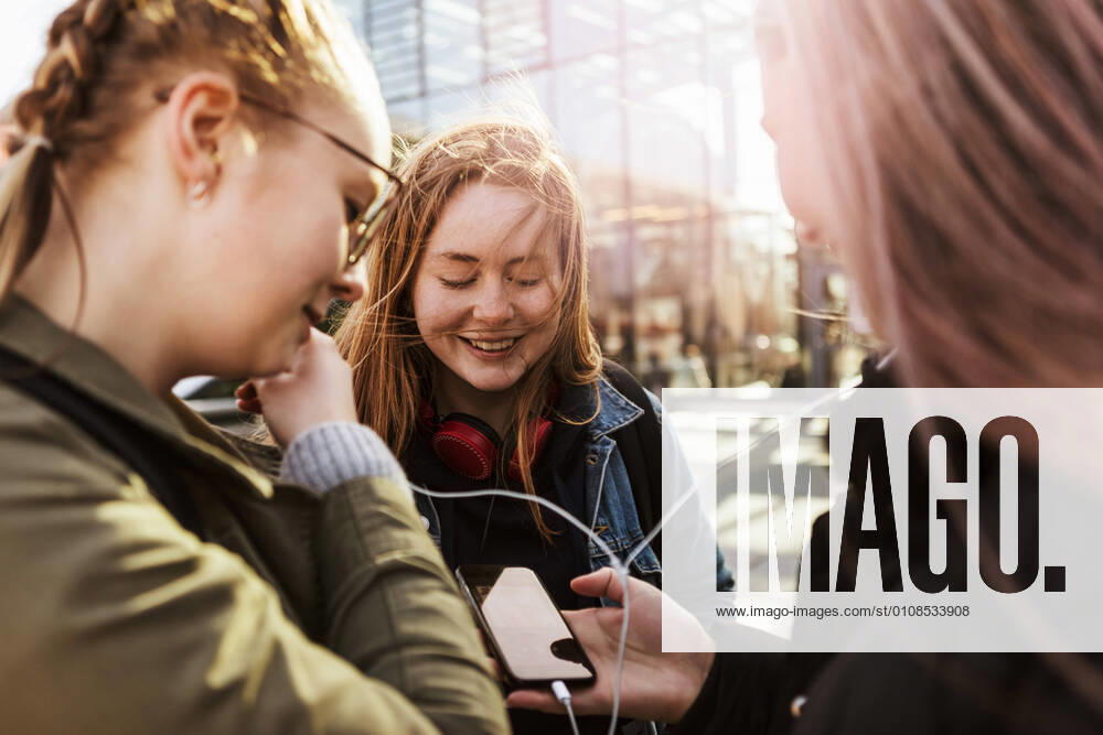 Teenage girl looking at friends listening music through mobile phone in city , model released