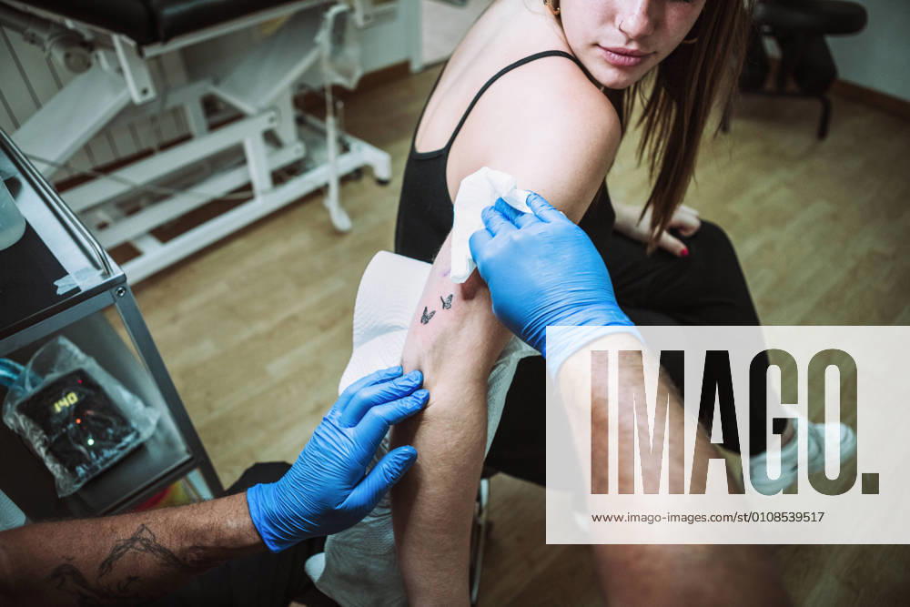 Back view crop anonymous male tattooist in latex gloves wiping freshly made tattoo on arm of female client with napkin while working in salon, Model released, Property released