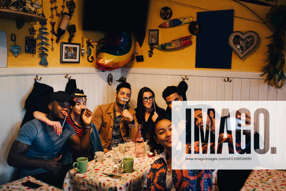 IMAGO Creative: Kimawandel - Weihnachten, Weihnachtsessen vegan Young woman taking selfie with cheerful multi-ethnic friends holding props while sitting at restaurant during dinner par , model released, property released