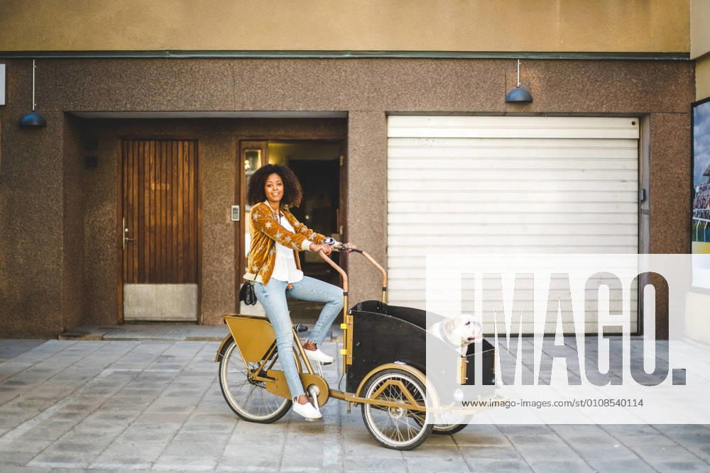 IMAGO Creative, Urban Lifestyle Full length of mid adult woman riding bicycle cart with dog against building , model released