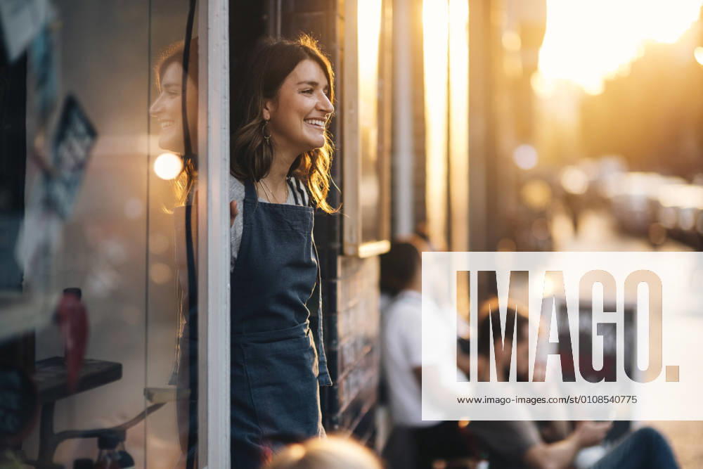 IMAGO Creative, Jahreszeit Sommer, Sommer in der Stadt IMAGO Creative, Frühling, Start der Straßencafe-Saison Smiling female owner looking away while standing at entrance of deli , model released, property released