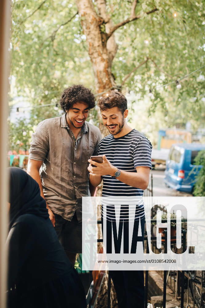 Cheerful young man showing smart phone to male friend while standing in balcony during party , model released, property released