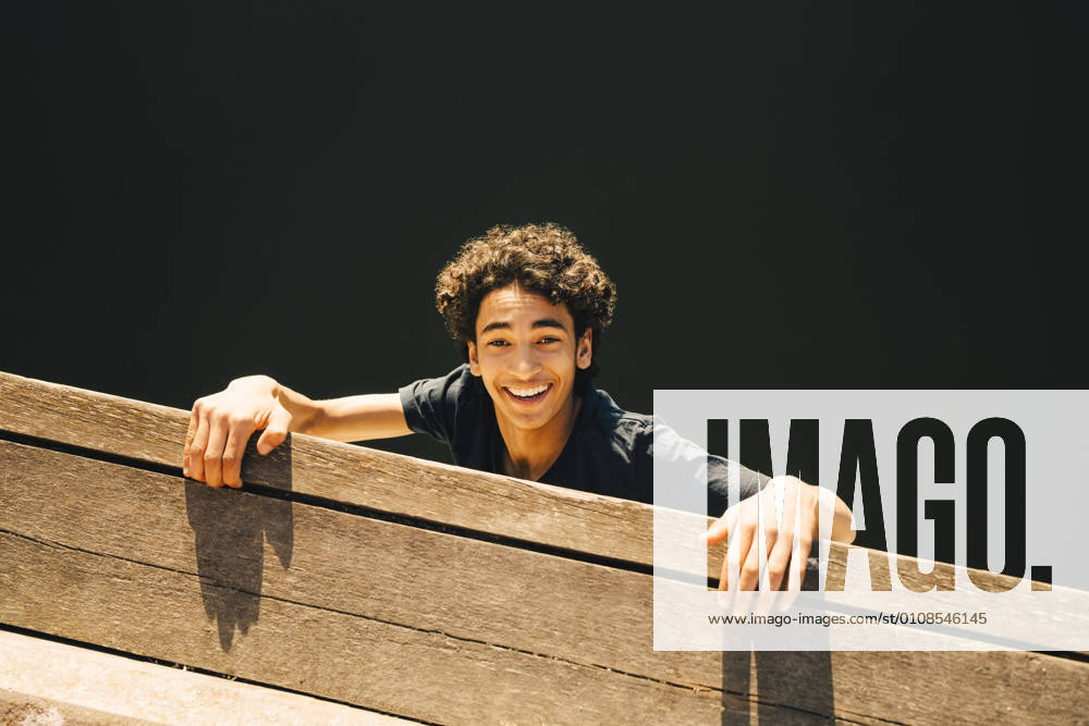 IMAGO Creative, Jahreszeit Sommer, Sommer in der Stadt High angle portrait of cheerful teenage boy hanging from pier against river , model released