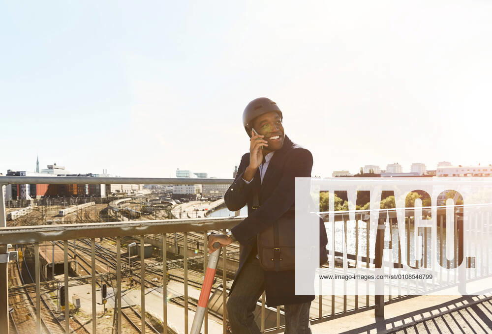 IMAGO Creative: Business, Innovative Technologie-Lösungen Smiling young man talking on smart phone while standing with electric push scooter at bridge in city against sky , model released