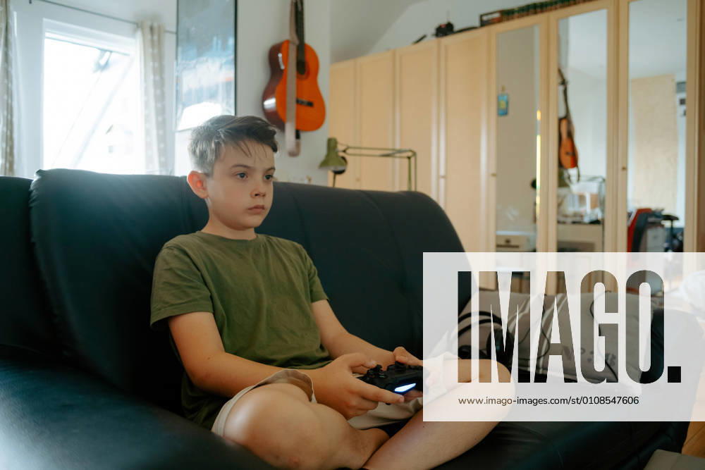 Concentrated little boy in casual clothes sitting on sofa with crossed legs and playing video game with joystick