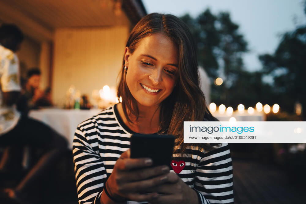 Smiling young woman using smart phone while friends in background during dinner party , model released, property released