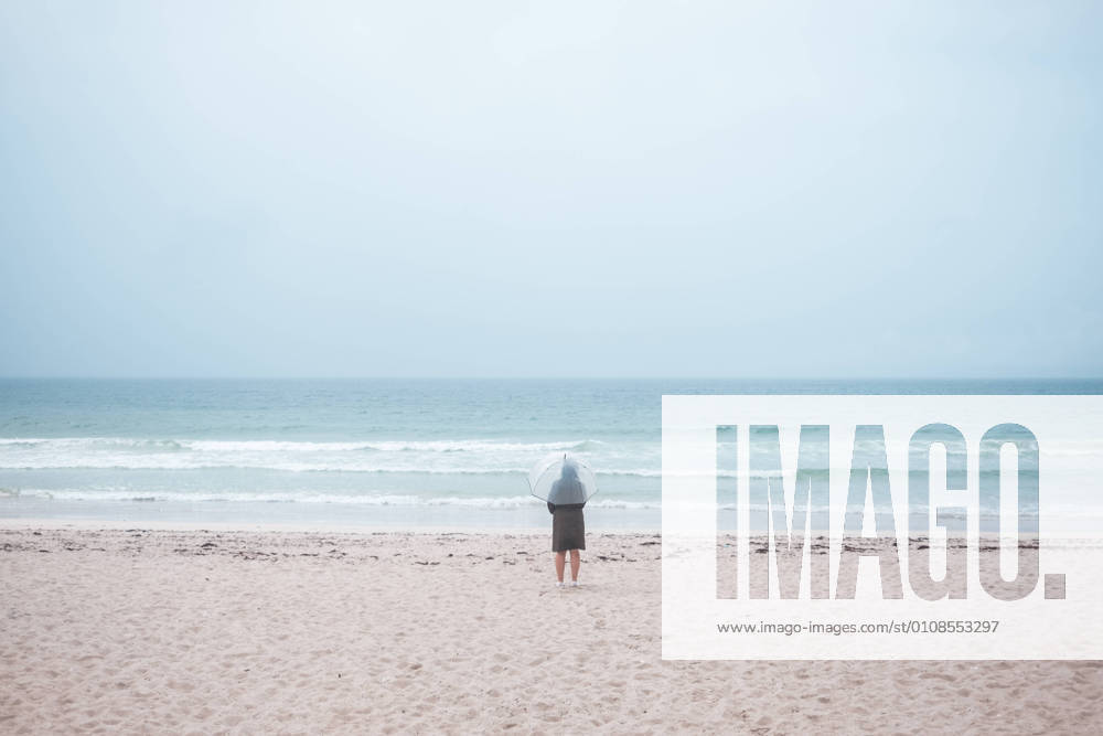 IMAGO Creative: Ende des Sommers Back view of unrecognizable traveler in raincoat with transparent umbrella walking on empty sandy beach towards waving sea in cloudy day