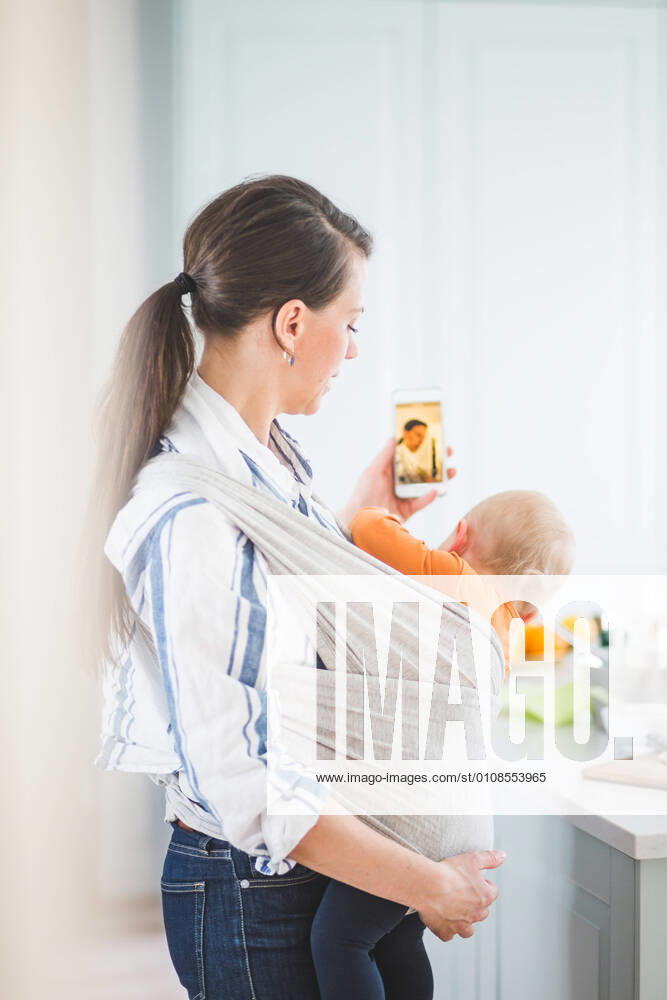 Side view of female blogger photo messaging while carrying daughter in kitchen at home , model released, property released