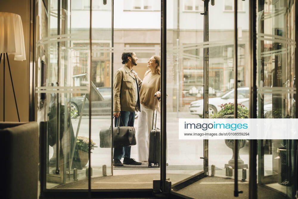 Couple with luggage talking while standing at doorway of hotel seen through glass , model released, property released
