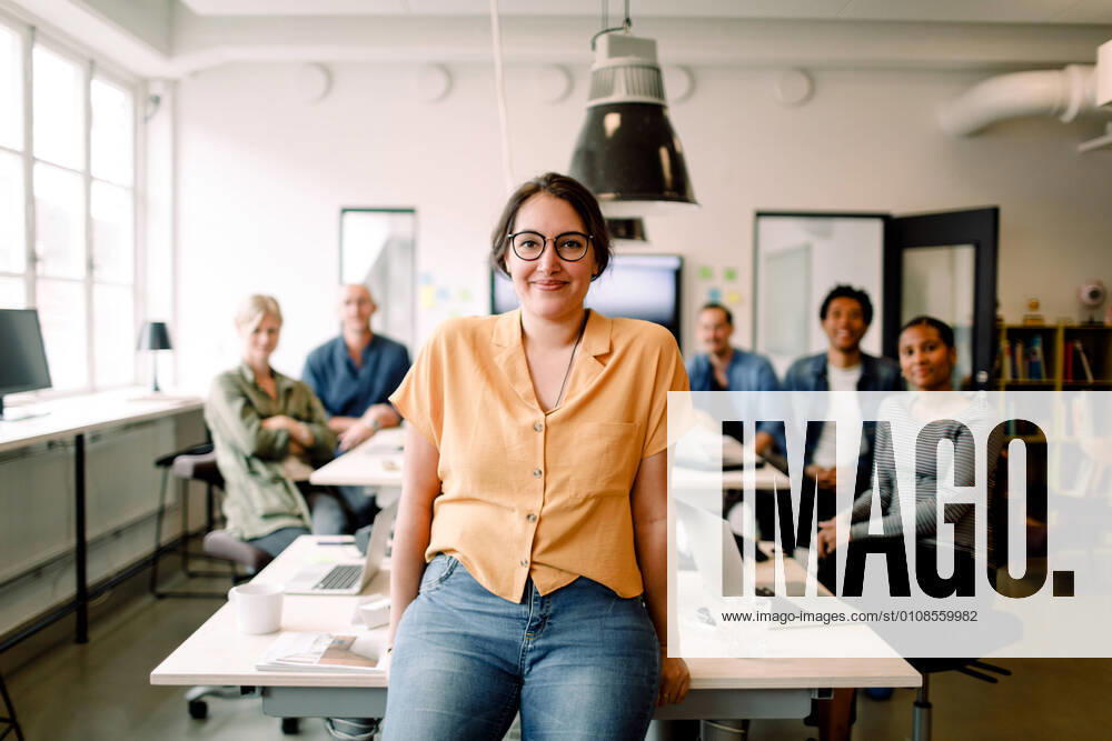 Portrait of smiling female professional leaning on table against executives in modern office , model released, property released
