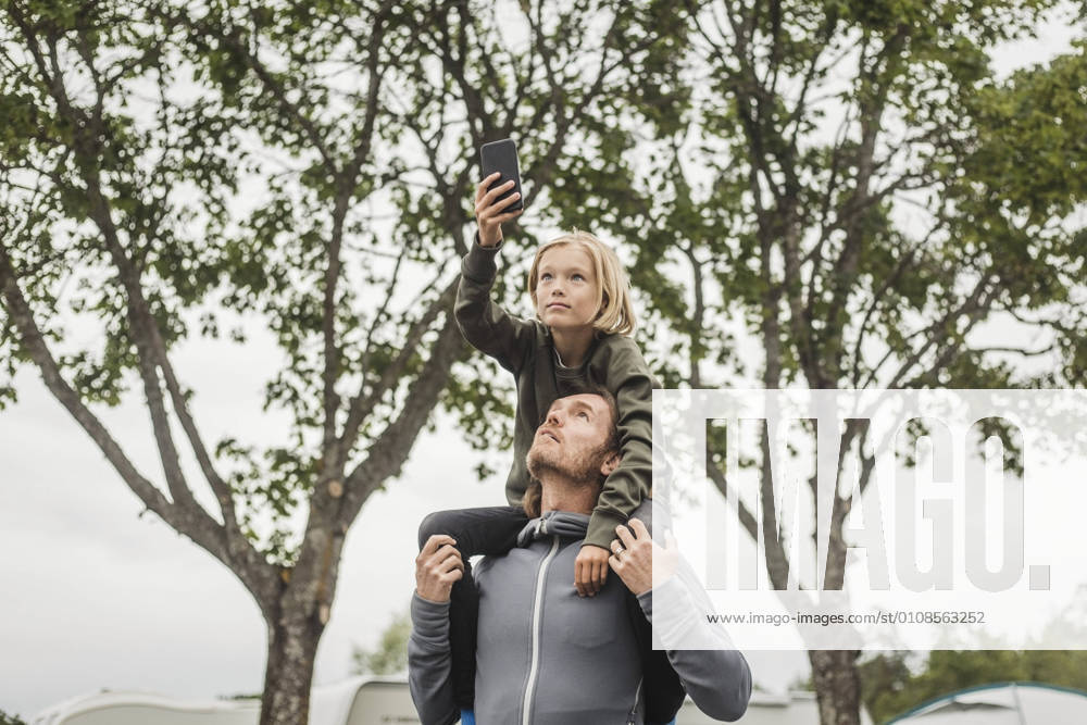 IMAGO Creative, Jahreszeit Sommer, Sommerferien Girl holding mobile phone while sitting on father s shoulders against trees , model released, property released