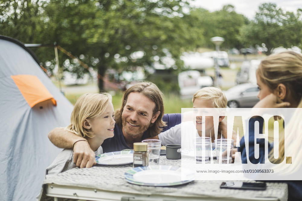 IMAGO Creative, Jahreszeit Sommer, Sommerferien Smiling father sitting with children at table in camping site , model released, property released