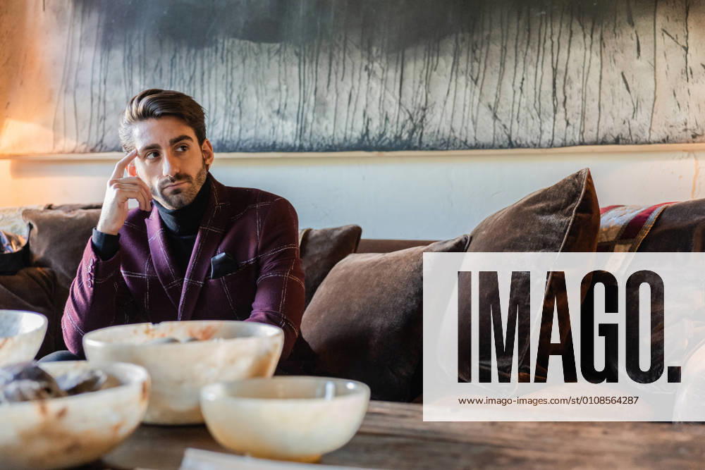 IMAGO Creative: Retro-Revival Pensive adult male in stylish clothes looking away and pondering while sitting on soft velour brown couch among cushions beside rustic wooden table against big gray modern picture in spacious apartment in retro style