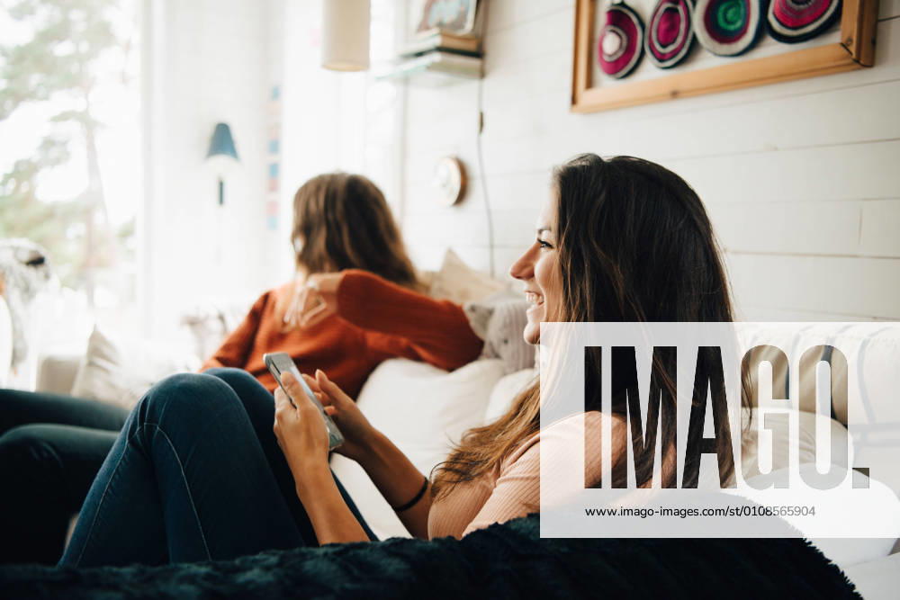 Smiling woman with smart phone looking away while sitting with friend on sofa at home , model released, property released