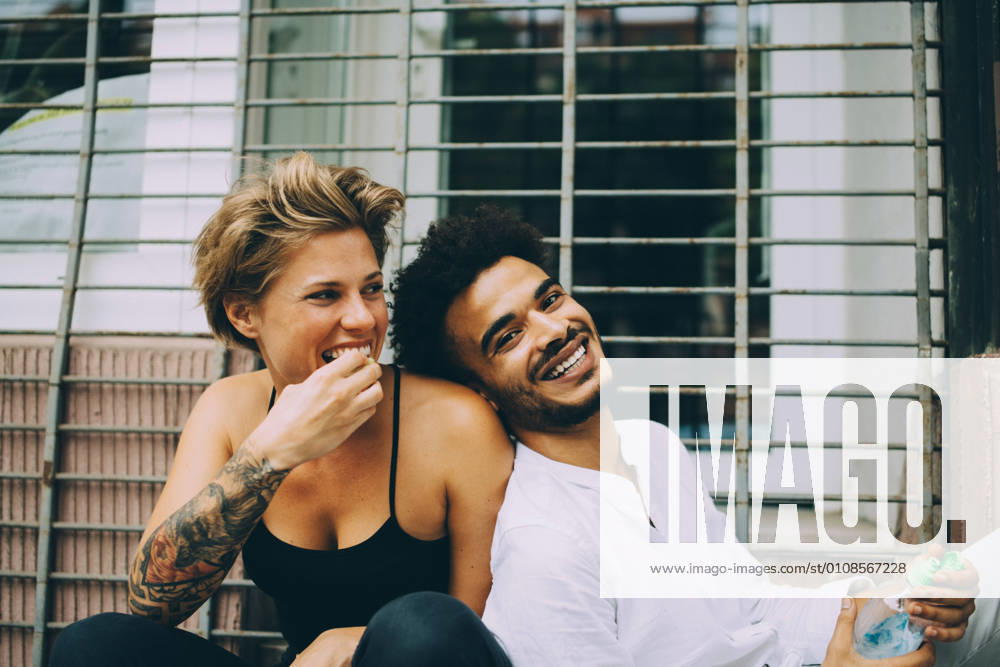 IMAGO Creative, Jahreszeit Sommer, Sommer in der Stadt Woman eating fruit while cheerful friend leaning on her against window , model released, property released