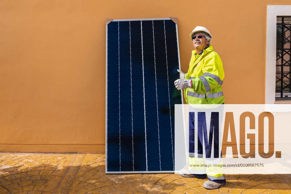 IMAGO Creative: Business, Innovative Technologie-L?sungen Side view of mature experienced male technician in uniform and helmet standing with solar panel near yellow building while working on installation of renewable energy system