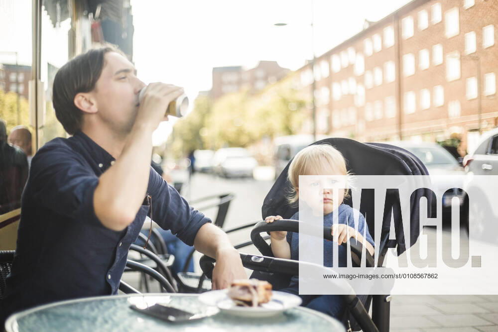 Baby boy looking away while sitting on stroller by father drinking at sidewalk cafe in city , model released