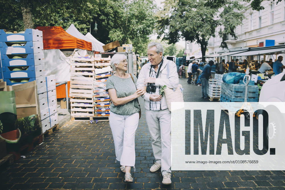 IMAGO Creative, Urban Lifestyle Full length of elderly couple talking while walking arm in arm at street market , model released