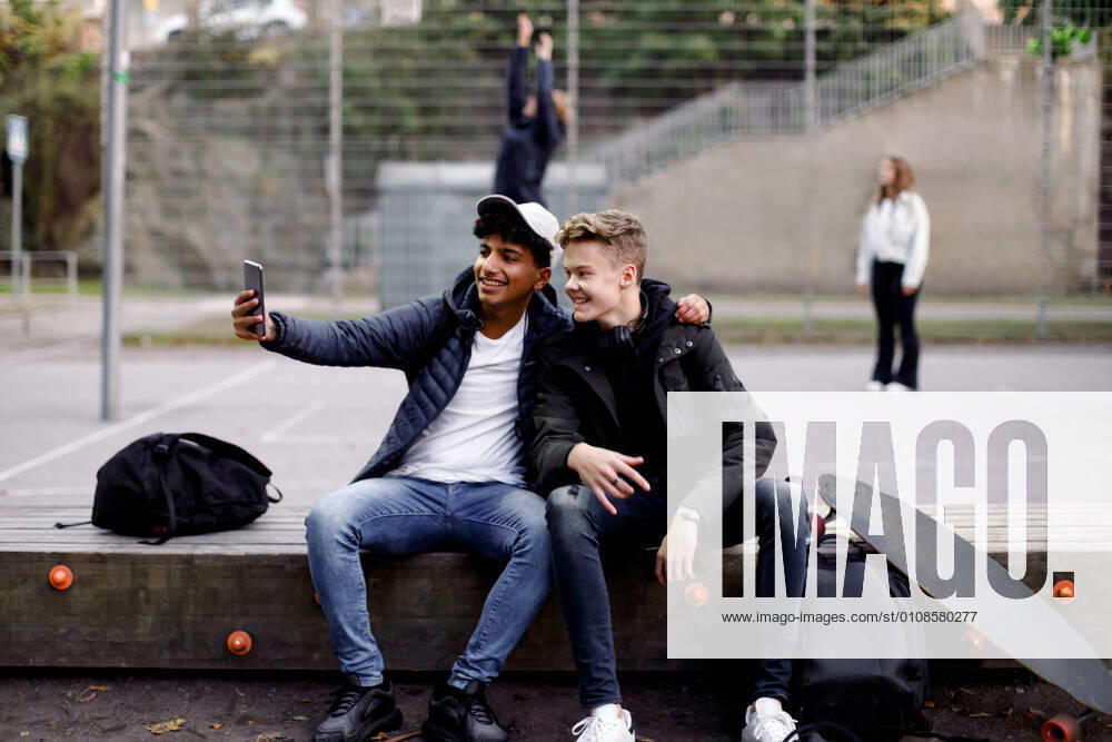 Full length of teenage boys taking selfie with mobile phone while sitting at park Sweden, Stockholm, model released