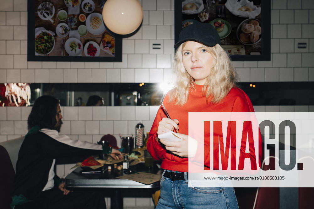 Portrait of female waitress with note pad standing against table in restaurant Sweden, Stockholm, model released, property released