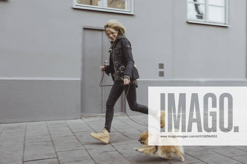 Full length side view of happy woman running with her Pomeranian dog on footpath by building in city Sweden, Stockholm, model released