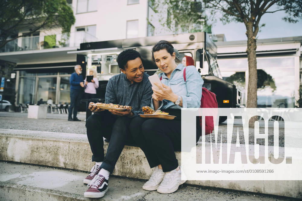 Smiling friends sharing smart phone while sitting with meal against food truck in city Sweden, Stockholm, model released, property released
