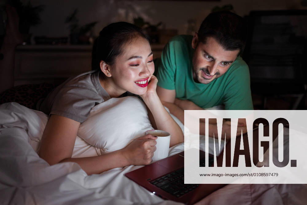 Smiling Asian female with cup of hot drink lying on bed near content boyfriend in casual wear while watching film on netbook at home