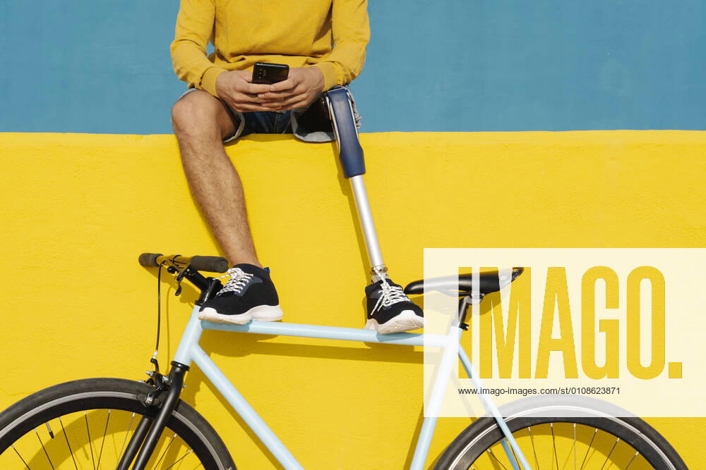 IMAGO Creative: Gesundheit, Prothese Young disabled man with bicycle using mobile phone while sitting on multi colored wall model released Symbolfoto JCZF00287