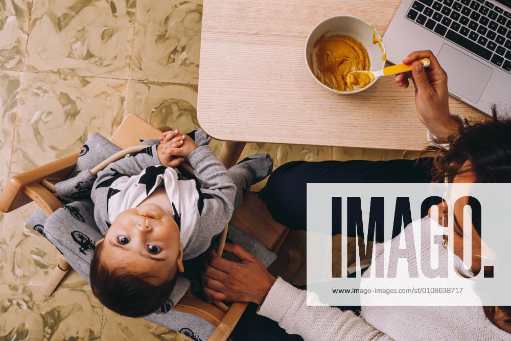 From above modern young female remote worker feeding little child while sitting at table with laptop and working on online project