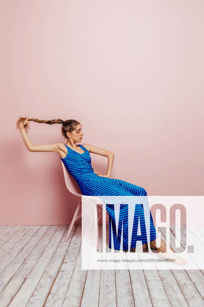 Side view of young woman in blue dress barefoot looking at camera holding ponytail while sitting in chair and looking away on minimalist pink background