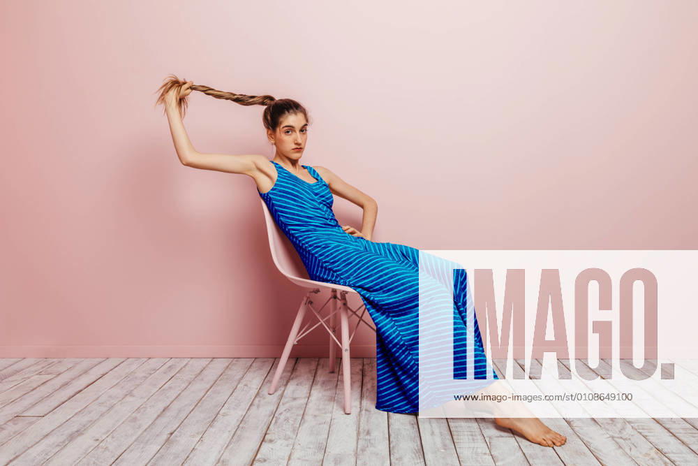 Side view of young woman in blue dress barefoot looking at camera holding ponytail while sitting with in chair and looking at camera on minimalist pink background