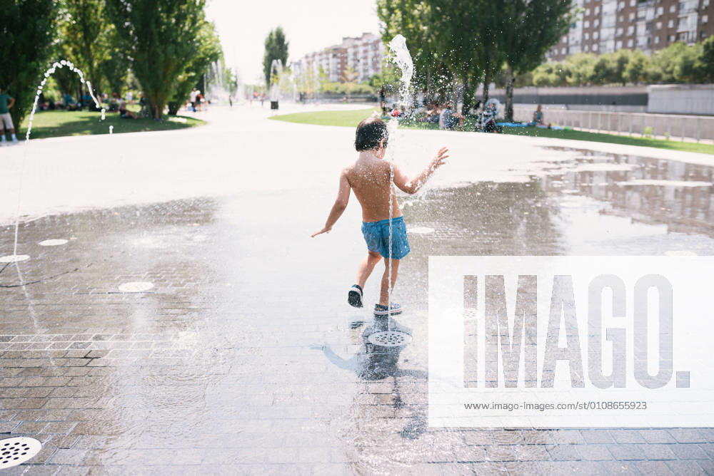 IMAGO Creative: Sommer, Sommertage im Stadtpark Back view of anonymous little boy in swimsuits standing near jet of water splashing out of fountain on street, Model released