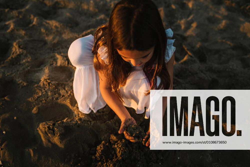 IMAGO Creative, Jahreszeit Sommer, Sommerferien Cute pensive female kid in white dress playing with sand on seaside in sunlight, Model released
