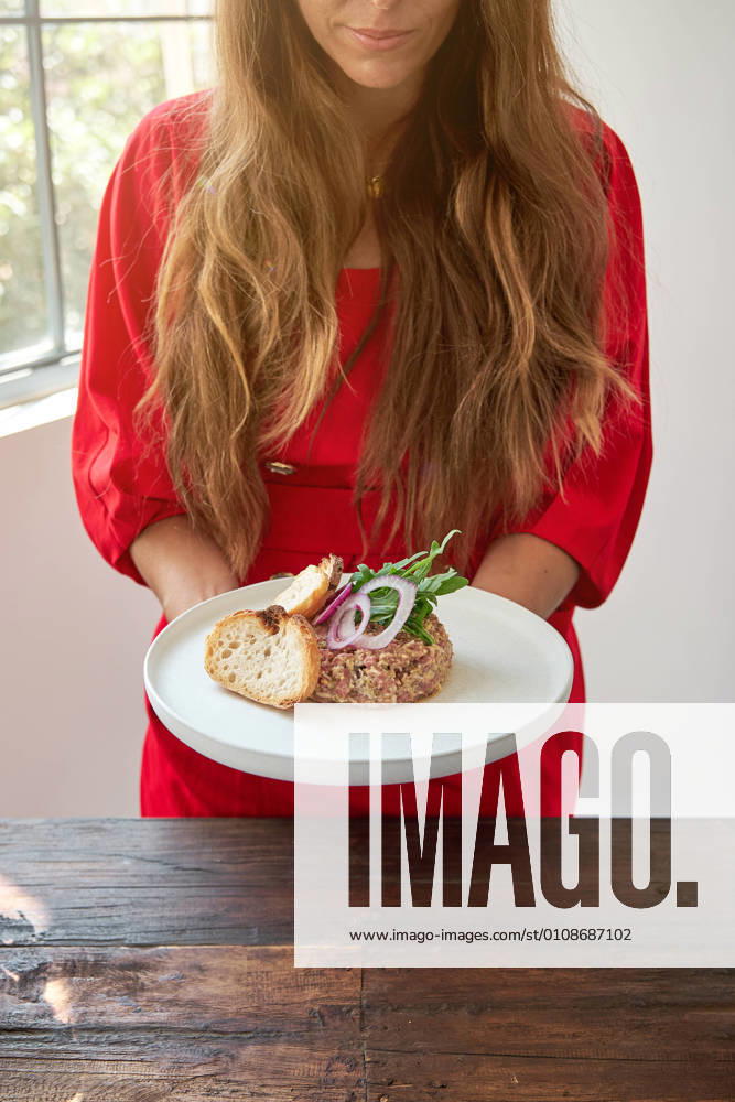 Crop unrecognizable female standing near wooden table with plate with appetizing Steak tartare garnished with fresh onion and rucola and served with bread, Model released