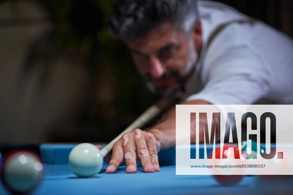 Confident focused gray haired male in white shirt preparing for shot with cue while playing billiards game